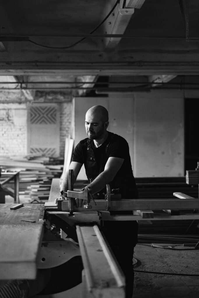 Black and white of skilled male carpenter cutting wooden detail at circular sawbench table while working in professional joinery with equipment